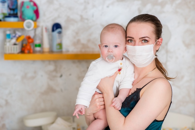 医療用防護マスクを着た母親が、コロノウイルスのパンデミックとcovid-19の最中に赤ちゃんを腕に抱えている