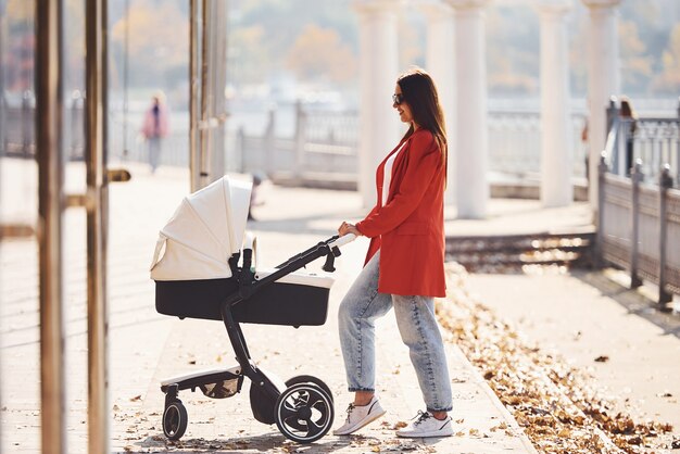写真 赤いコートを着たお母さんは、秋の公園の乳母車で子供と散歩をしています。