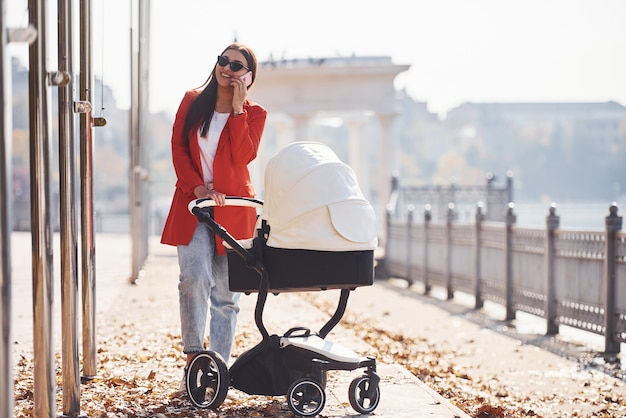 写真 赤いコートを着たお母さんは、秋の公園の乳母車で子供と散歩をしています。