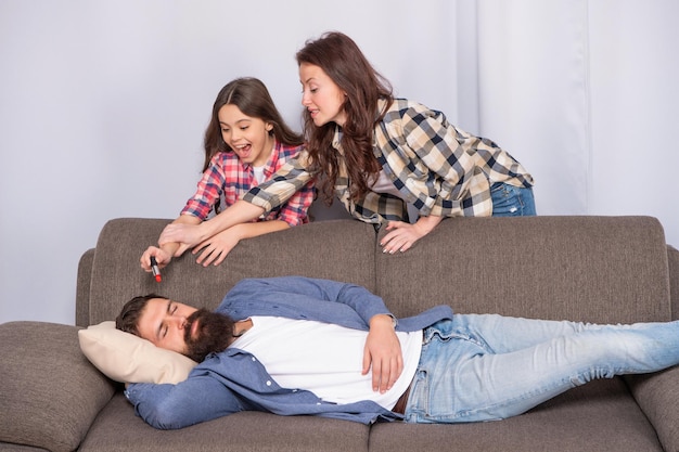 Мать удерживает девочку от нанесения помады отцу, спящему на диване дома