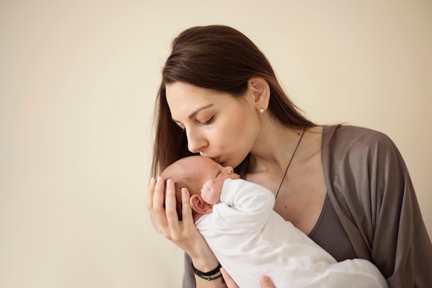 Mother kissing newborn son at grey background. Portrait of woman and little baby love