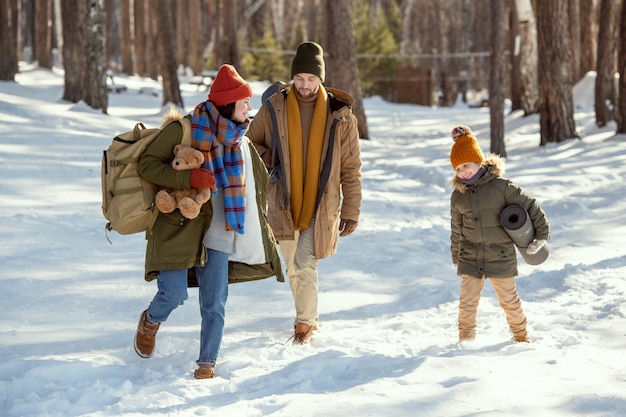 Мать и дочка в теплой зимней одежде веселятся в сугробе по пути в загородный дом, отец следует за ними в зимний день