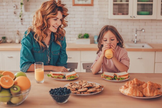 Madre che guarda bambina che beve succo d'arancia mentre fanno colazione insieme