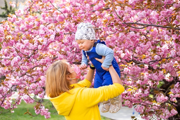 公園で幼い息子と一緒に医療マスクの母は桜の木の近くを歩く