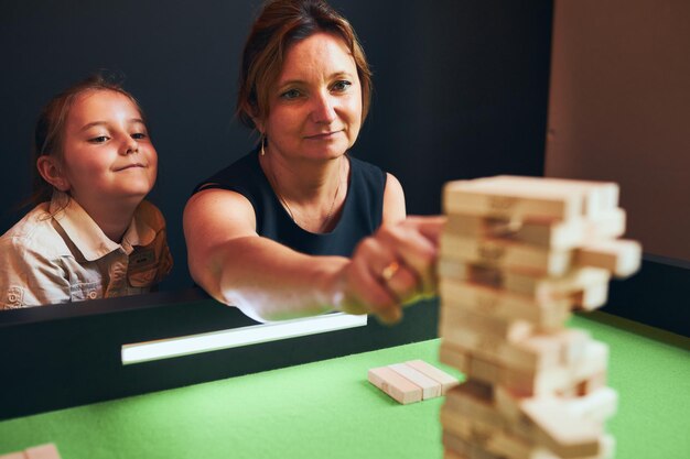 Photo mother playing jenga game with her daughter in play room woman removing block from stack game room