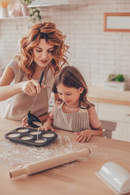 Mother pouring batter with ladle into the form for muffins and daughter helping her on the kitchen