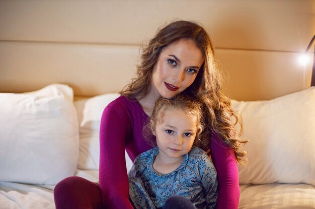 Mother in purple clothes with a sad dissatisfied daughter in dress is sitting on a bed in a hotel