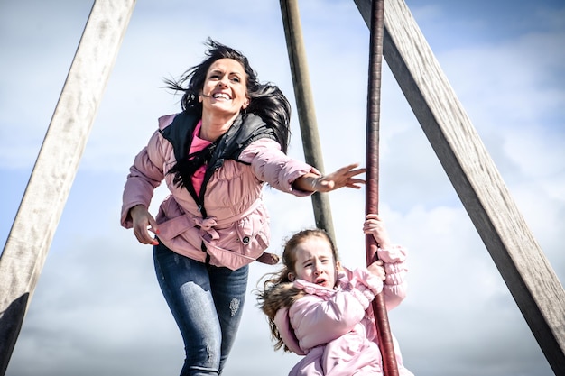 Mother pushing daughter sitting on swing in playground against sky