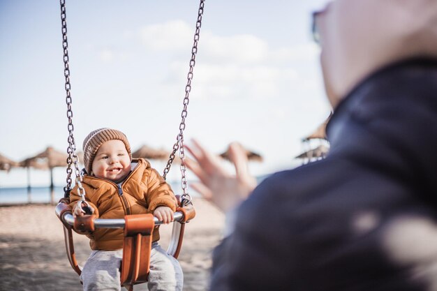 Mother pushing her cheerful infant baby boy child on a swing on sandy beach playground outdoors on nice sunny cold winter day in Malaga Spain