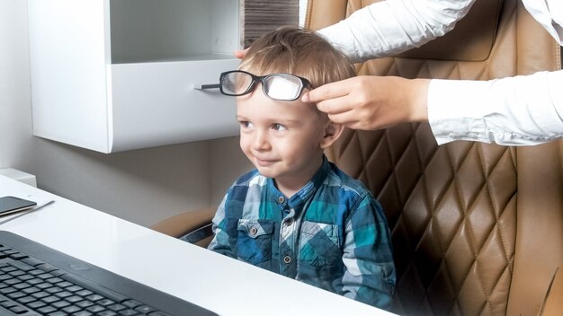 Mother putting eyeglasses on her little toddler son sitting in office chair.