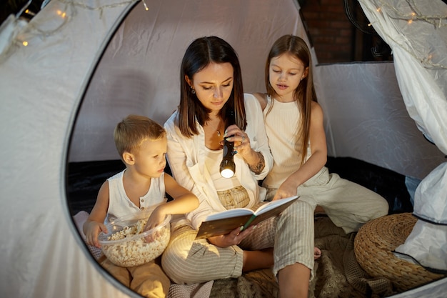 Mother reads a book of fairy tales for her children while sitting in a tent at night. Mom son and daughter reading a book with a flashlight in their hands