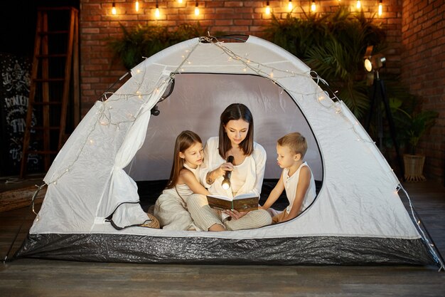 Mother reads a book of fairy tales for her children while sitting in a tent at night