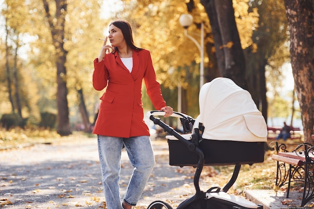 Mother in red coat have a walk with her kid in the pram in the park at autumn time and smoking.