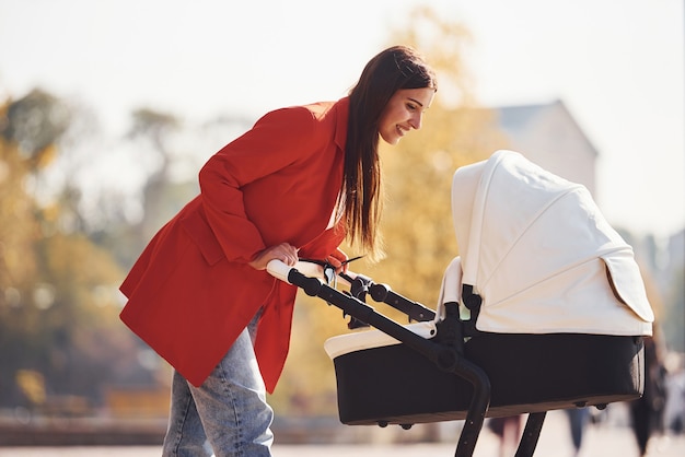 Mother in red coat have a walk with her kid in the pram in the park at autumn time.