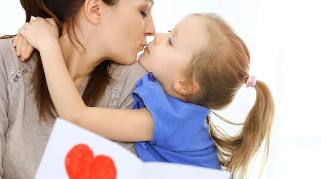 Mother's day concept. Child daughter congratulates mom and gives her postcard with red heart shape. Mum and girl happy smiling and hugging. Family fun and holiday.