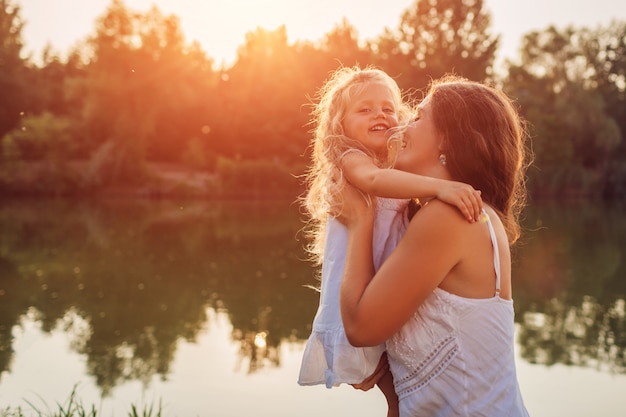 Mother's day. Woman playing having fun with daughter by summer river at sunset. Woman holding kid and laughing. Family