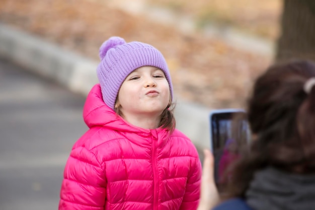 秋の公園で彼女のかわいいポーズの小さな娘を携帯電話のカメラで撮影する母