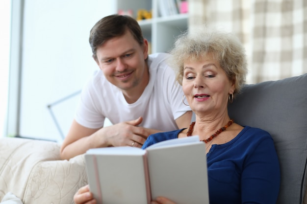 Mother sit on couch, hold photo album and show her son photo.