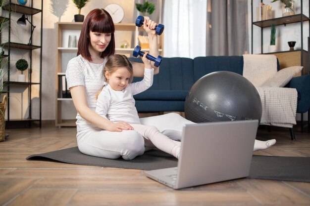 Mother sitting on mat with little charming daughter in sportswear doing exercises with dumbbells