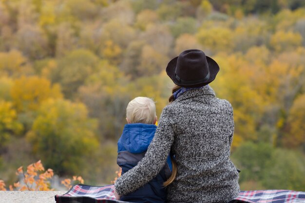 Mother and son are sitting on a high ground. Autumn. Forest in the distance. Back view