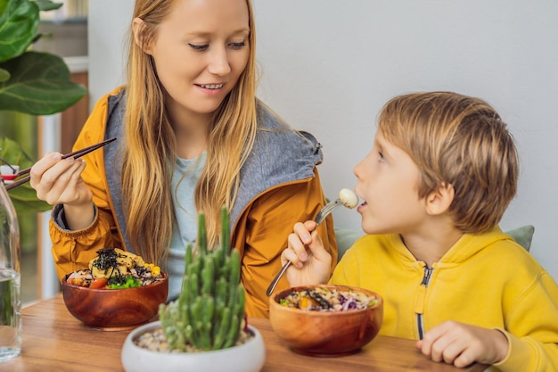 Mother and son eating raw organic poke bowl with rice and veggies closeup on the table top view from