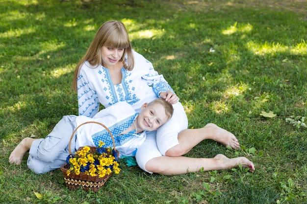 A mother and son sit on the grass with a basket of flowers.