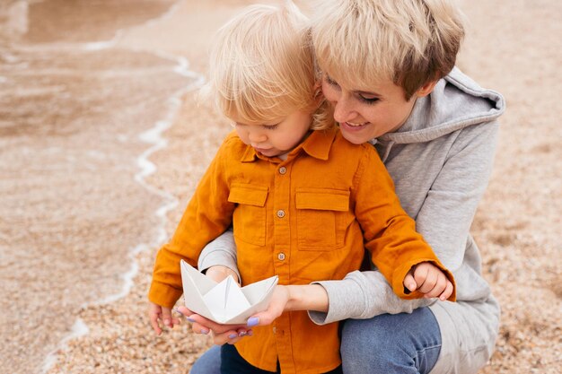 Mother and son sitting on the beach playing with paper boat in autumn time