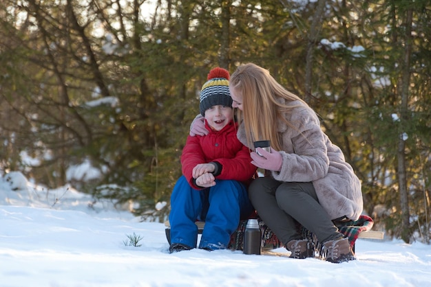Mother and son sitting on a sled in the winter forest chatting and drinking tea from a thermos