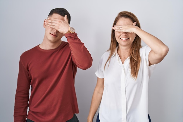 Mother and son standing together over isolated background covering eyes with hand looking serious and sad sightless hiding and rejection concept