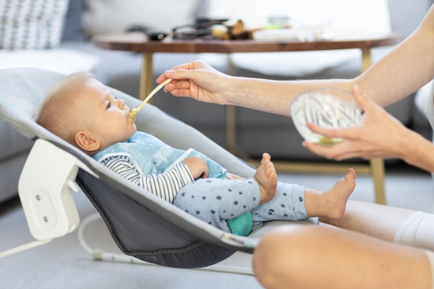 Mother spoon feeding her baby boy child in baby chair with fruit puree baby solid food introduction