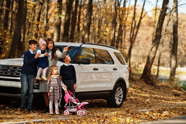 Foto la madre sta vicino all'auto suv bianca con quattro bambini nel parco autunnale. passeggiata in famiglia nella foresta autunnale.