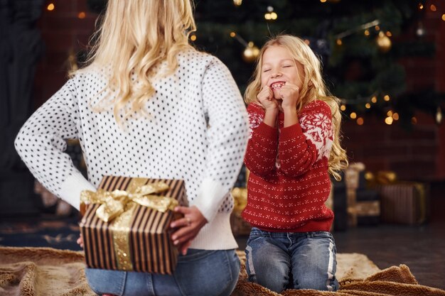 ギフトボックスを持って立っている母親は、クリスマス休暇の時間に屋内で娘を驚かせます。