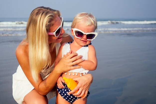 Mother in stylish glasses and white dress is putting orange spray bottle SPF on newborn baby one year old daughter a sunscreen on the beach