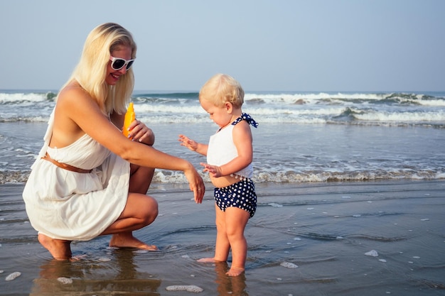 Mother in stylish glasses and white dress is putting orange spray bottle SPF on newborn baby one year old daughter a sunscreen on the beach