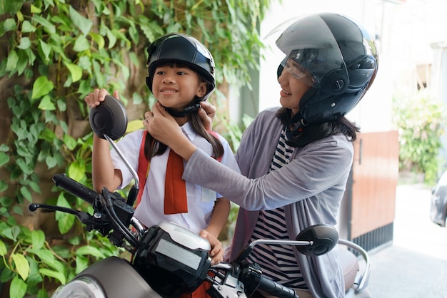 朝、バイクで娘を学校に連れて行く母親。制服を着たアジアの小学生が学校に戻る