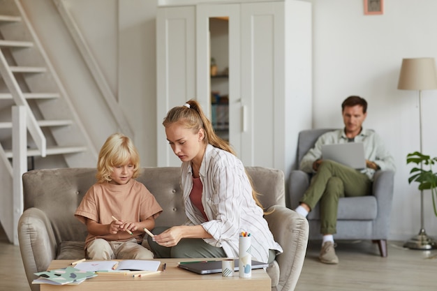 Mother teaching her son at the table in the room with father working on laptop 