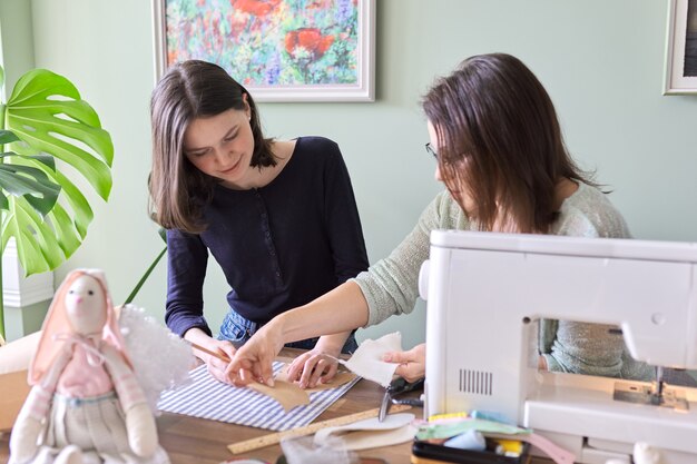 Mother and teen daughter sew together at home. Mom teaching daughter sewing on machine, women sew toy and clothes for toy. Parent and teenager, relationships, lifestyle, creativity, skill