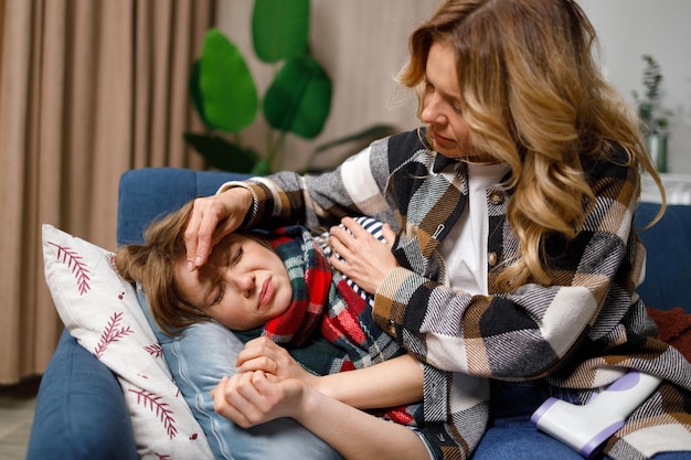 Mother touches the forehead of her daughter who is lying under the blanket on the couch