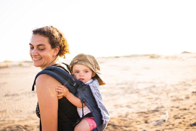 Mother turning to see the baby that holds on her back in a baby carrier outdoors