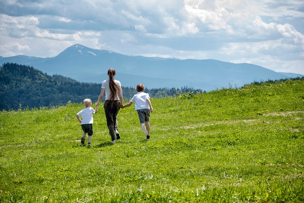 Mother and two young sons running on green field holding hands in green mountains and sky with clouds.