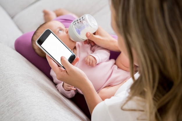 Мать с помощью мобильного телефона во время кормления ребенка бутылкой молока