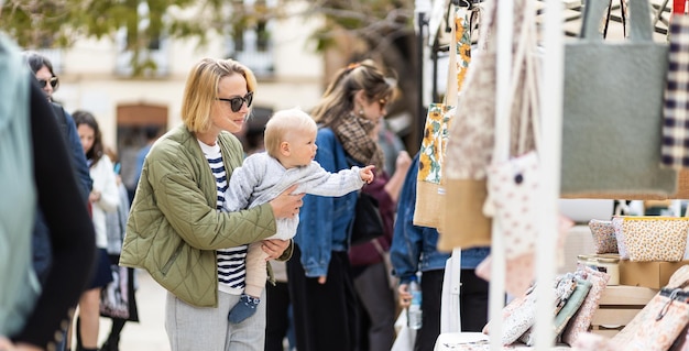 Mother walking carrying his infant baby boy child in crowd of people wisiting sunday flea market in