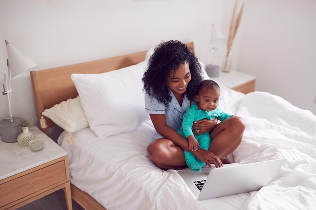 Мать в пижаме с маленькой дочерью в спальне работает из дома на ноутбуке