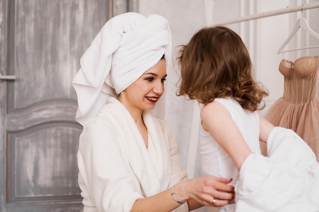 mother in a white bathrobe is dressing her little daughter