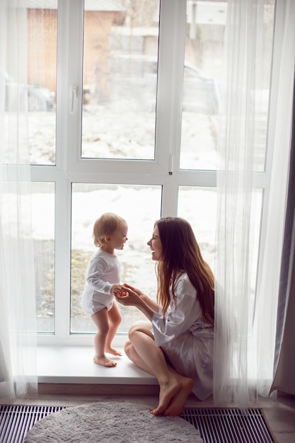 Foto madre in una vestaglia bianca si siede con un bambino una figlia bionda ad una grande finestra della casa persona