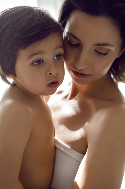 Mother in a white t-shirt holding her son in her arms standing at the window