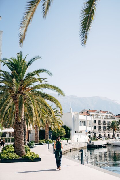 Mother with a baby in a sling walks along the embankment past palm trees back view