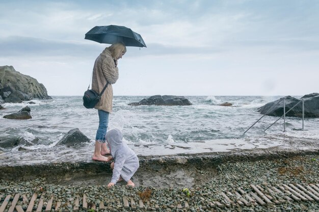 悪天候のビーチで石の多いビーチで子供と小さな男の子と母海の海岸沿いを歩く