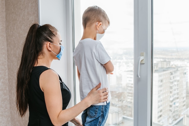 Mother with a child in medical masks are sitting at home in quarantine and look out the window. Prevention of Coronovirus and Covid -19