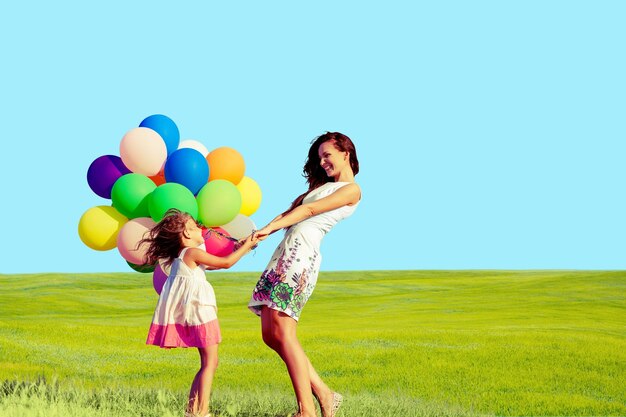 Mother with daughter holding colorful balloons on grass and sky background